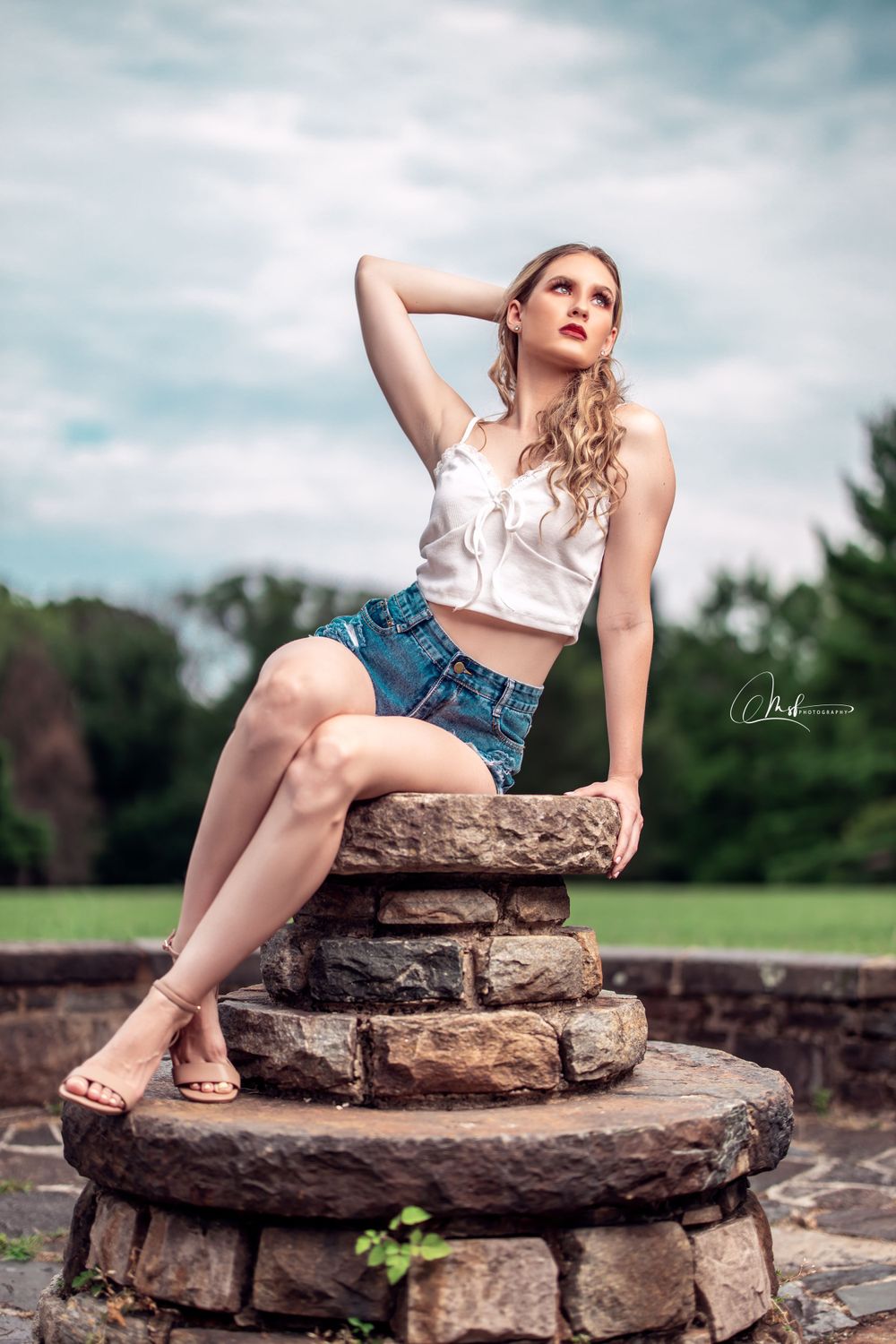Model in white crop top and denim shorts poses on stone structure in outdoor garden setting.