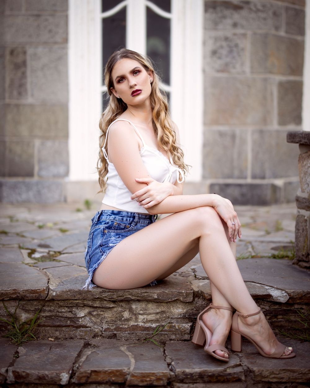 Woman in white top and denim shorts seated on stone wall with classic architecture backdrop.