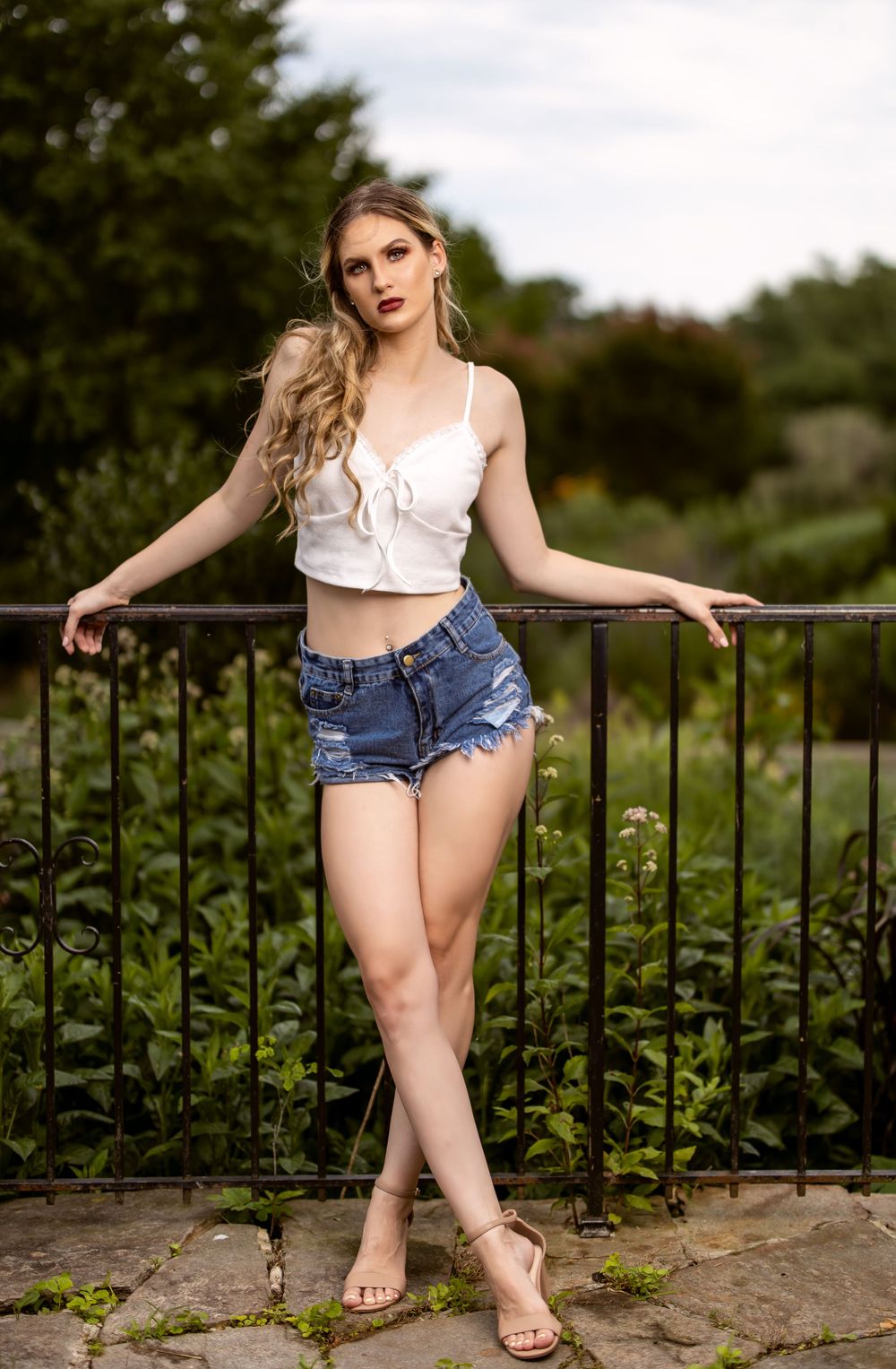 Fashion portrait of woman in summer outfit leaning against iron railing in garden.