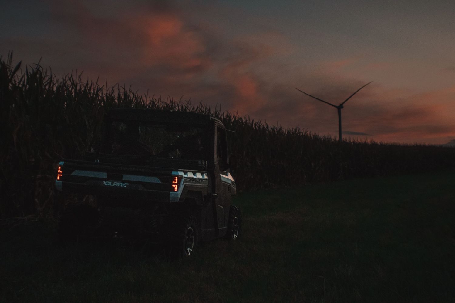 Silhouette of a vehicle against darkening sky with wind turbine and cornfield in background.