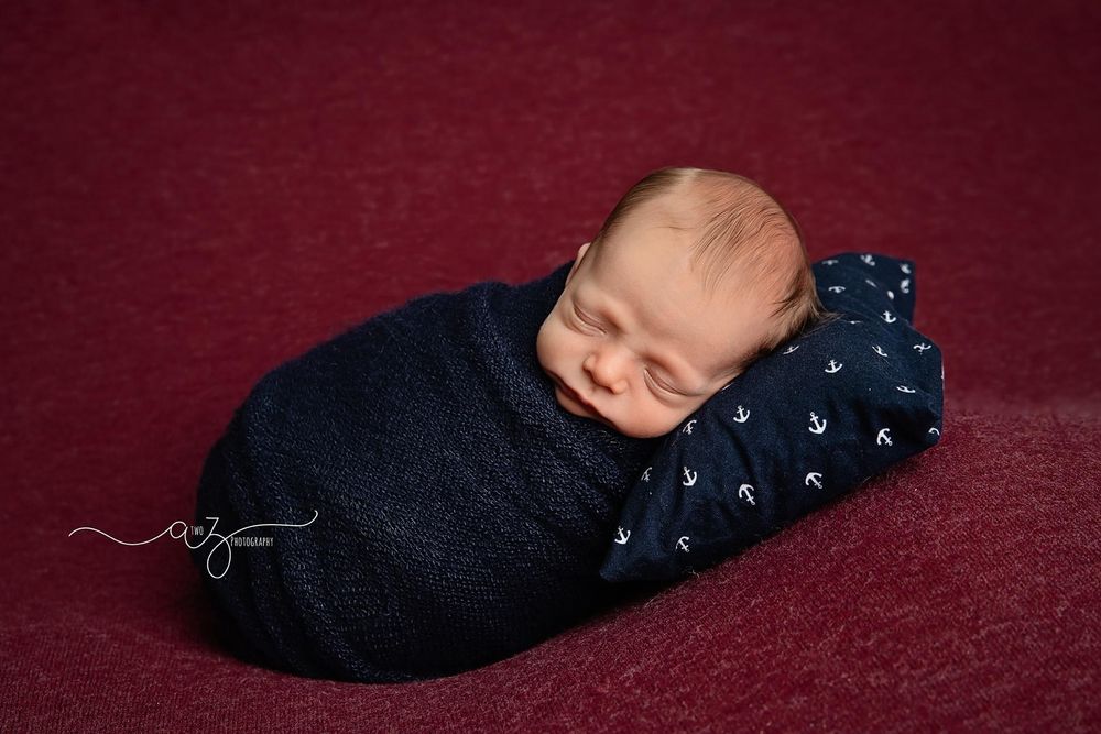 Sleeping newborn wrapped in navy swaddle on nautical print pillow against burgundy backdrop.