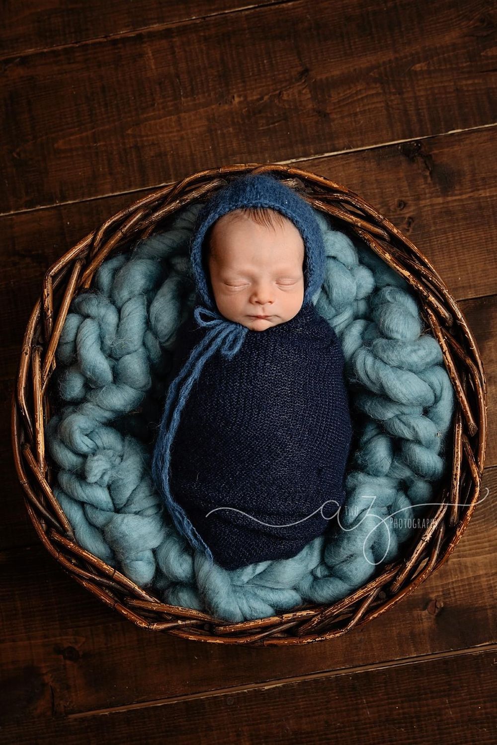 Sleeping newborn wrapped in navy knit on teal chunky knit blanket inside rustic woven basket.