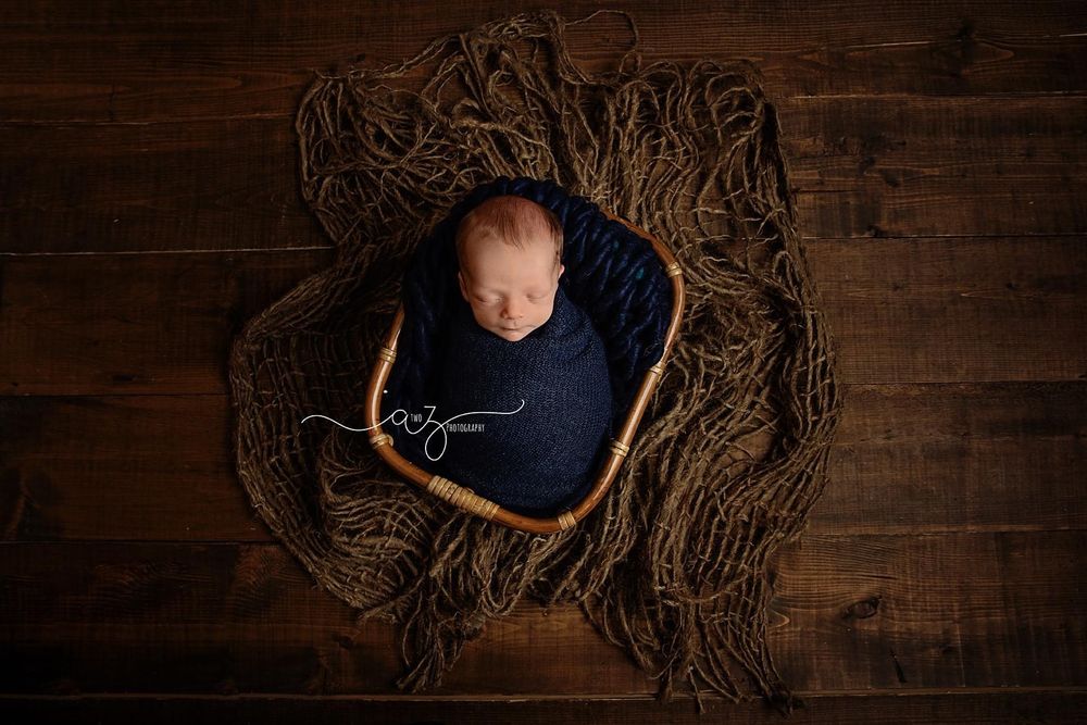 Overhead view of sleeping newborn in navy wrap nestled in wooden bowl with textured backdrop.