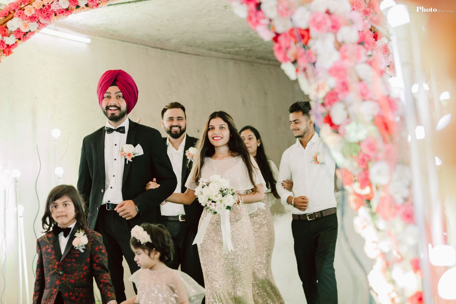 Wedding party walks under pink floral arch with festive lighting.