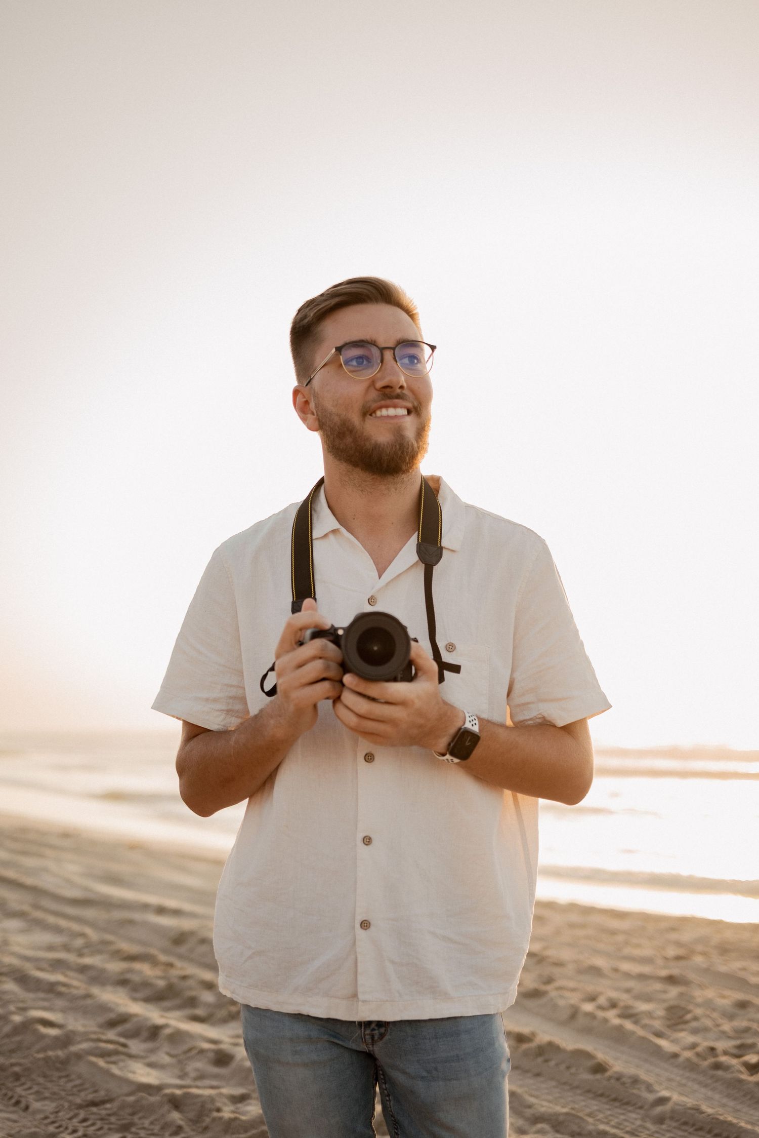 A photographer with a camera stands on a beach at sunset wearing a white button-down shirt.