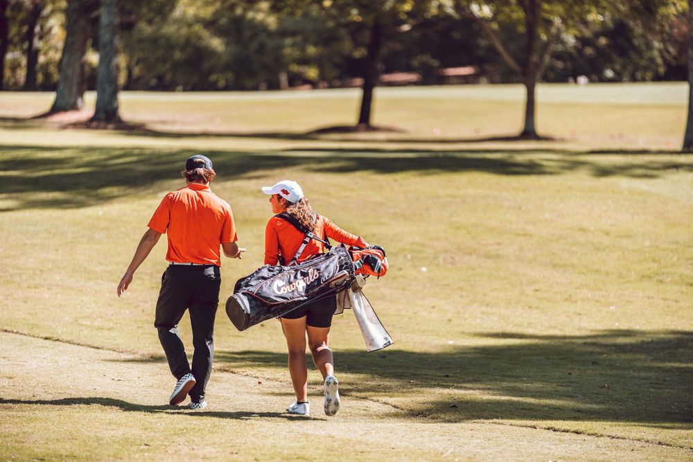 Two golf players walking on a sunny course with one carrying clubs and golf bags.