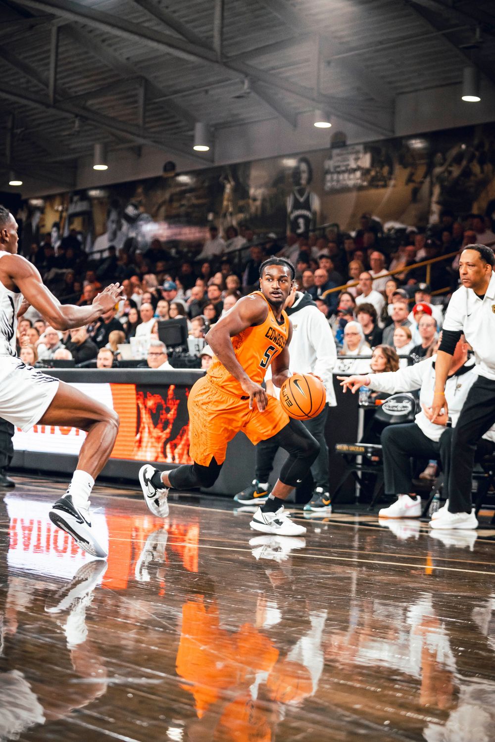 Player in orange jersey drives past defender during competitive basketball game on shiny hardwood floor.