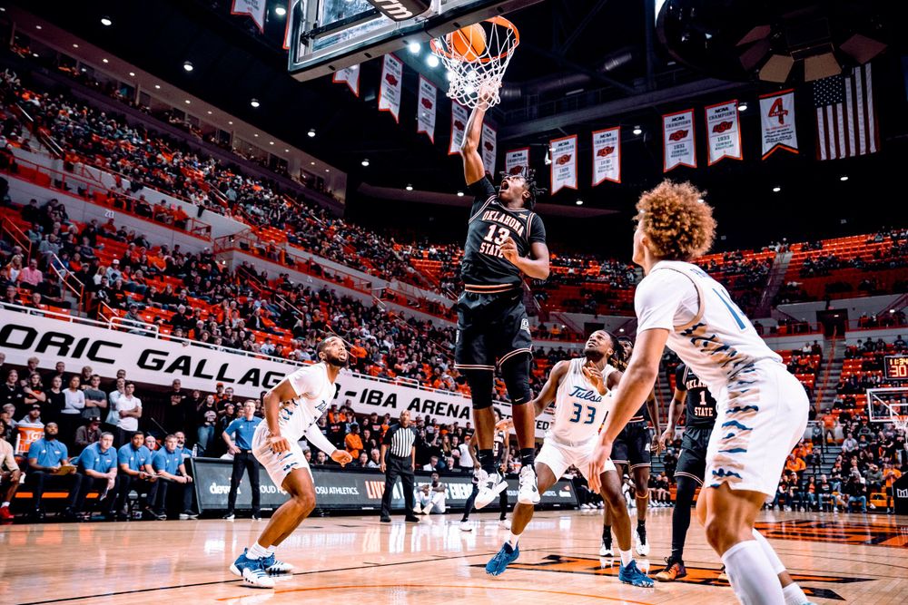 Basketball player rises up for a shot in a packed arena with banners and flags visible.