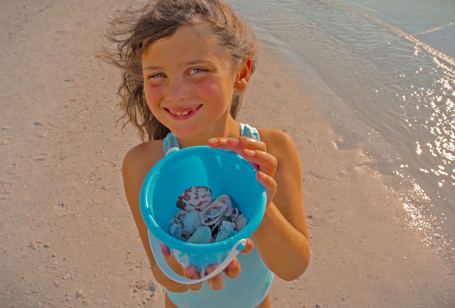 Smiling girl holding a teal bucket of seashells on a sunny beach.
