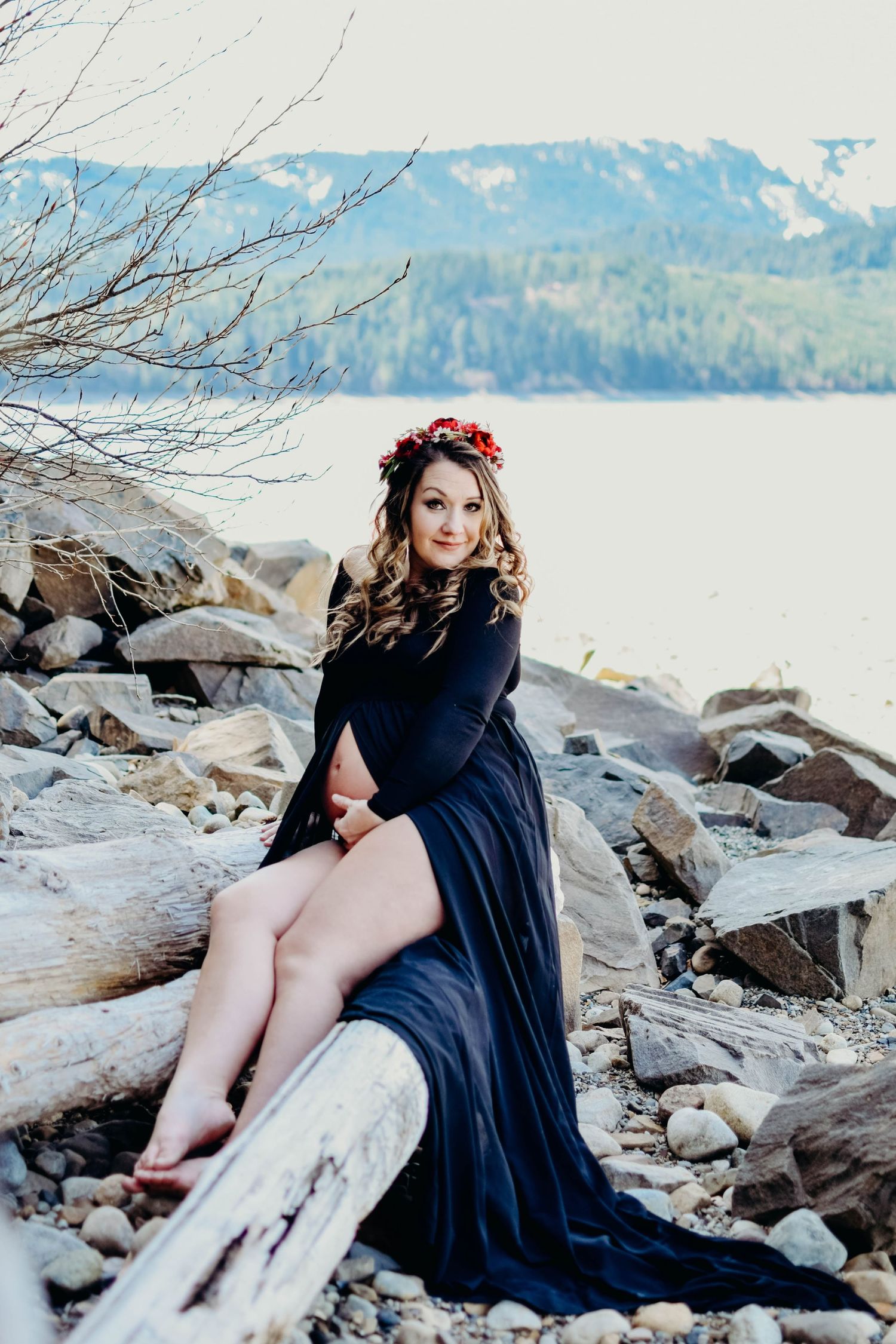 Woman in black dress posing on rocky shoreline with lake and mountains in background.