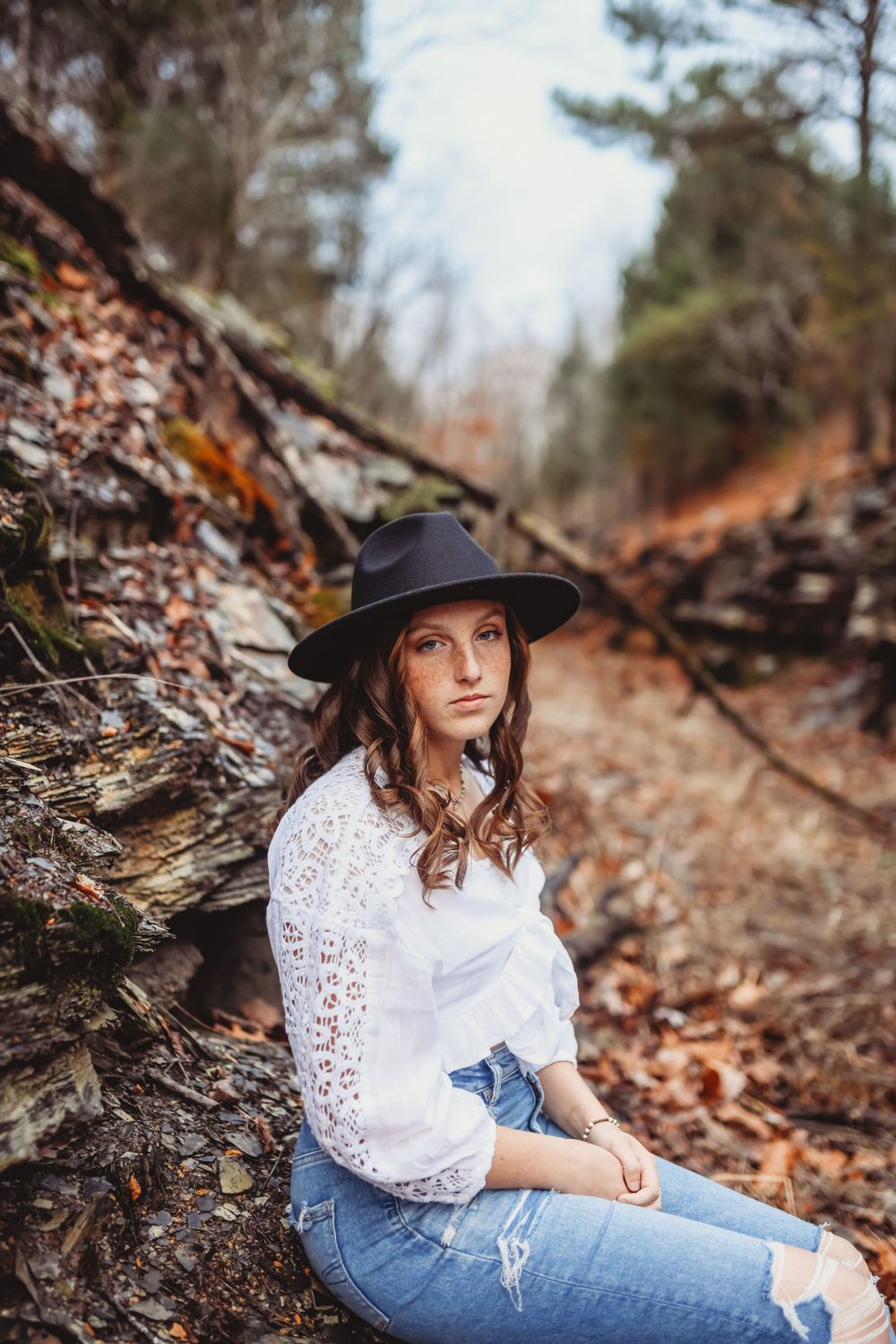 Autumn forest photoshoot featuring white lace top and wide-brimmed hat among fallen leaves.