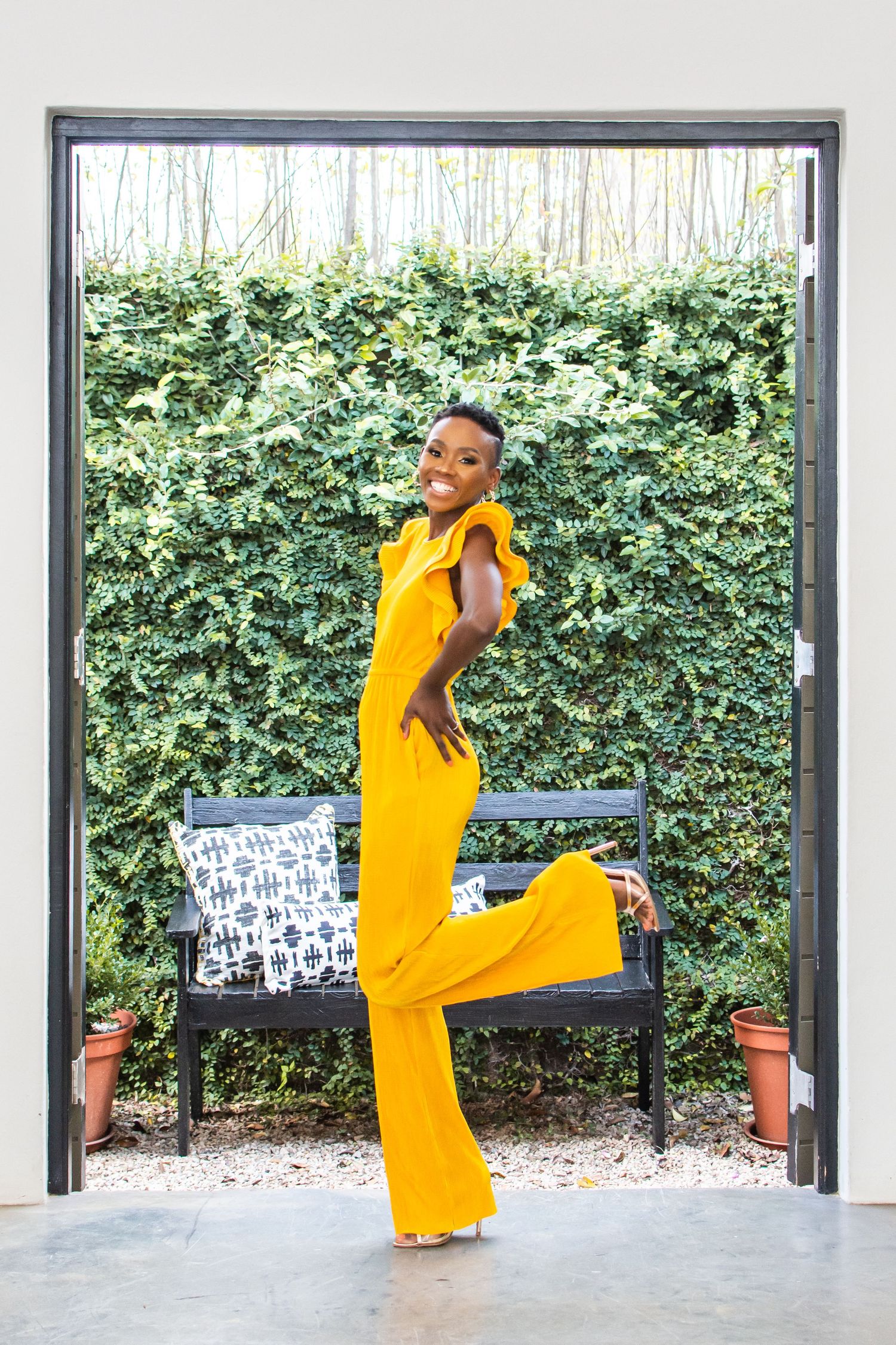 Full-length yellow jumpsuit fashion photo posed in doorway with garden background.