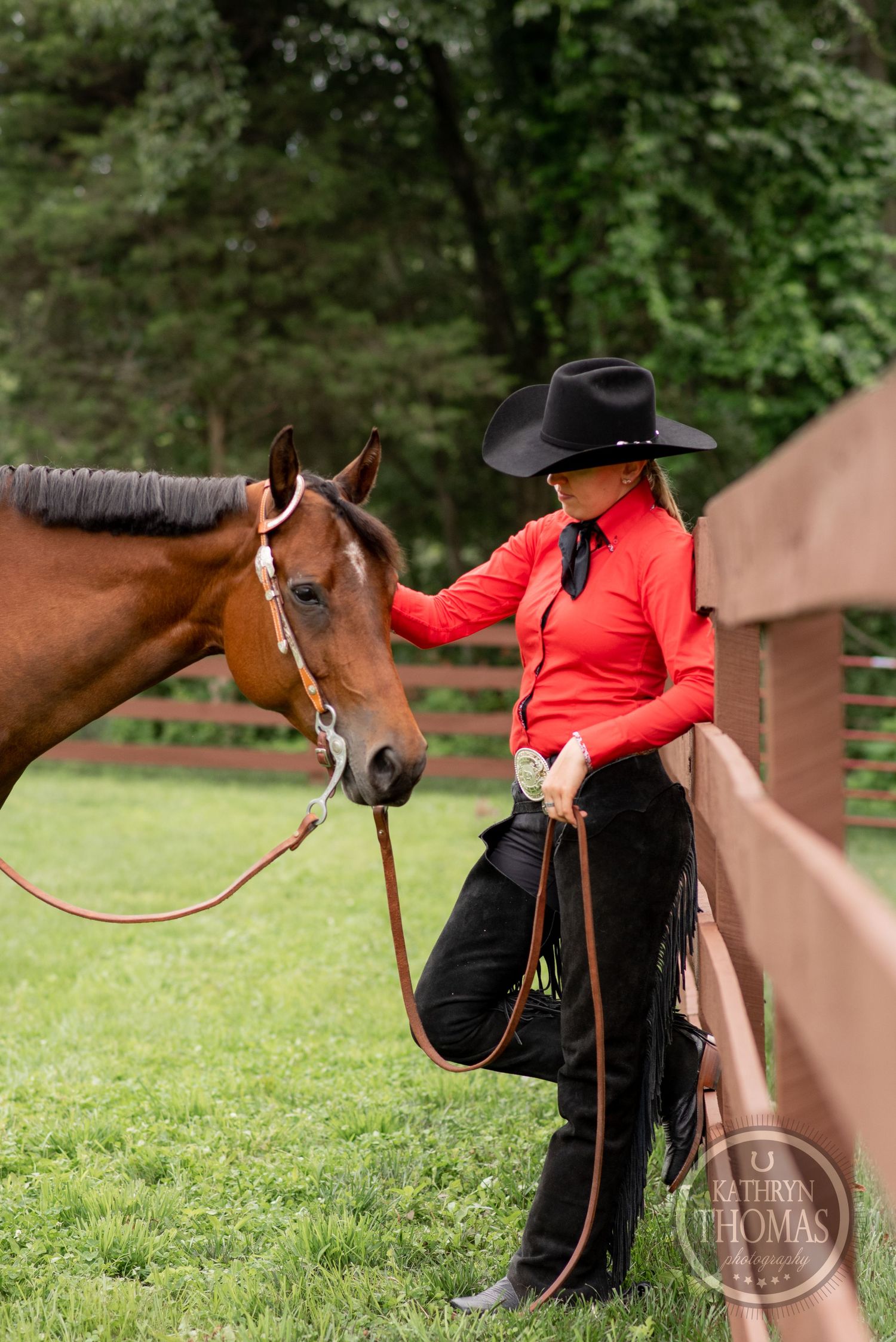 Equestrian in red western shirt and cowboy hat poses with horse near wooden fence.