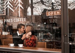 Cozy coffee shop scene through window showing couple sharing intimate moment at cafe table.