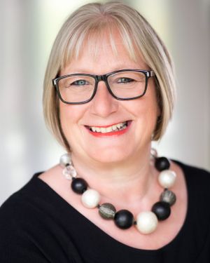 Professional portrait in black top with black and white statement necklace and glasses.
