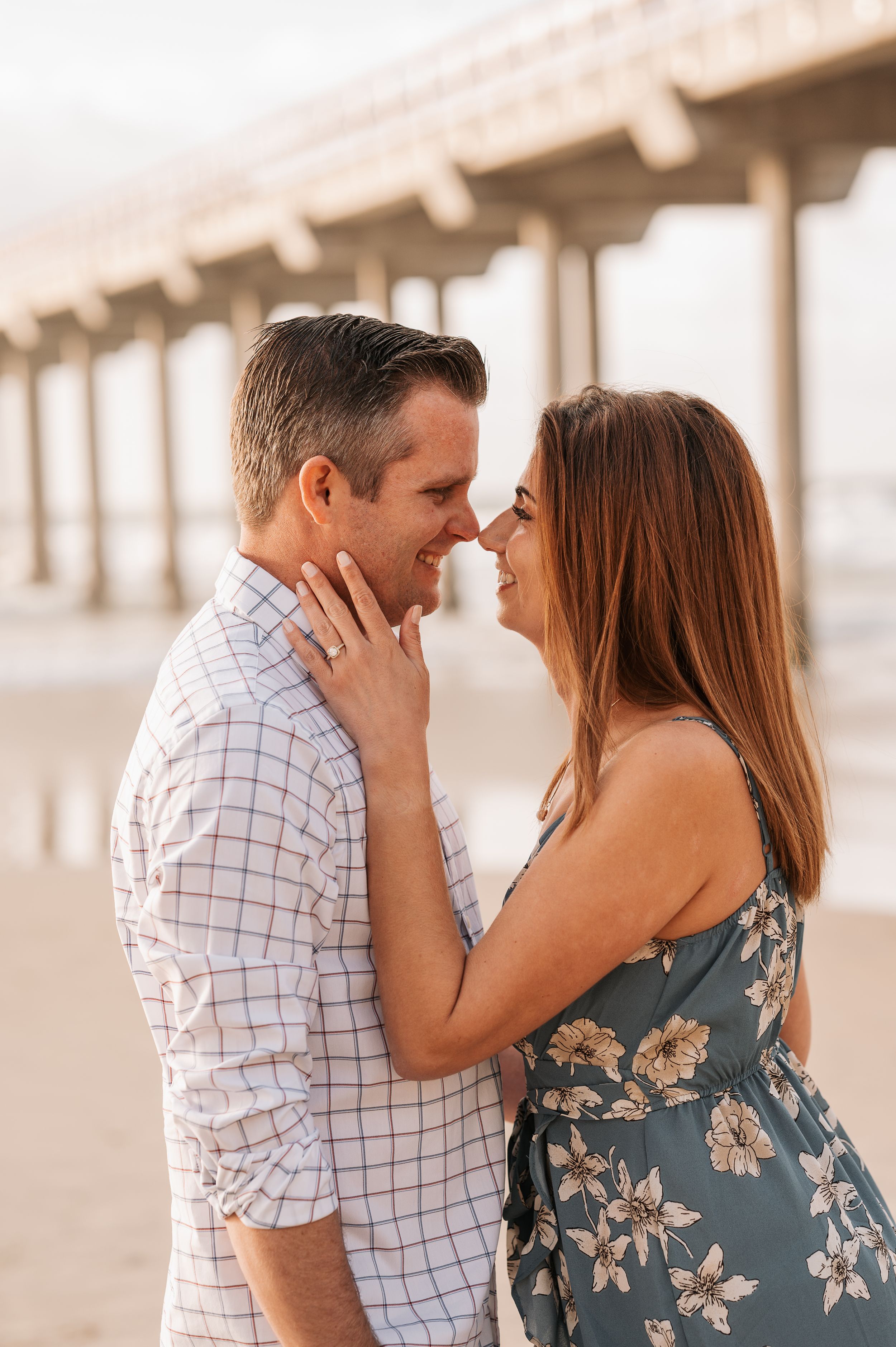 Intimate moment between couple in casual attire on beach with pier in background.