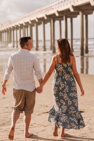 A couple walks hand in hand beneath a wooden pier on the beach, with waves rolling in the background.