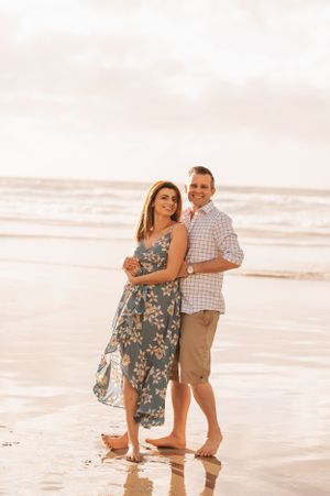 A romantic couple poses barefoot on a sandy beach at sunset, with her in a floral dress and him in khaki shorts.
