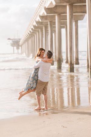 A couple shares a joyful embrace under a pier with ocean waves reflecting on wet sand.
