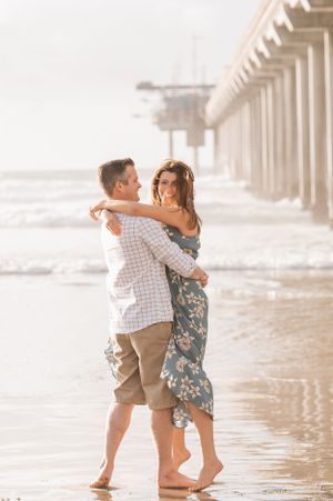 A couple embraces in shallow water beneath a pier during a romantic beach photoshoot.