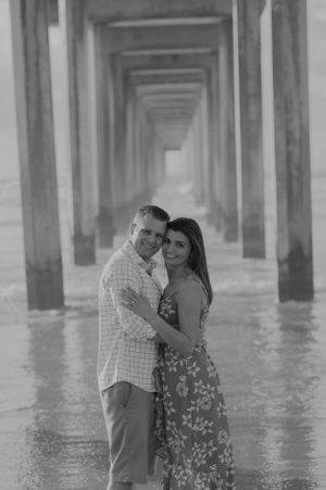 A black and white portrait of a couple embracing under a pier's geometric columns.