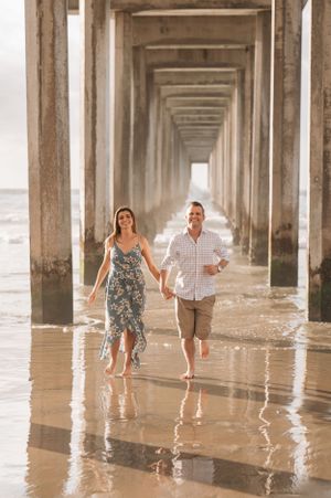 A couple runs through shallow water beneath a pier during golden hour.
