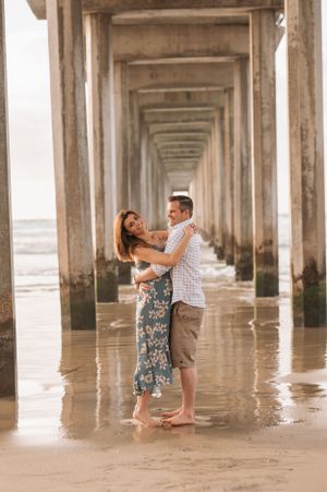 A couple dances beneath a pier's columns during a romantic beach photoshoot.