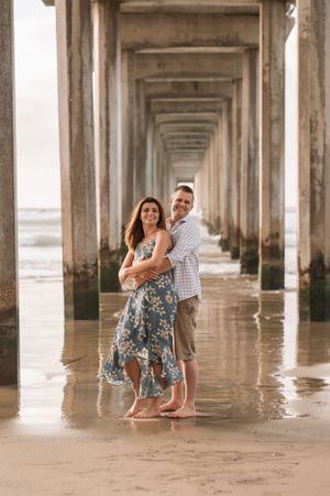 A couple poses between symmetrical pier columns with ocean waves in the background.