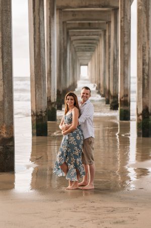 A couple poses beneath a beach pier in floral dress and casual wear with ocean waves in the background.
