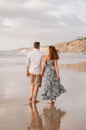 Couple walking barefoot on wet sand beach with cliffs in background during sunset.