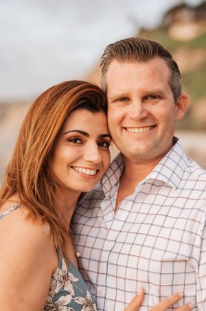 Couple in casual attire smiling at beach with coastal background.