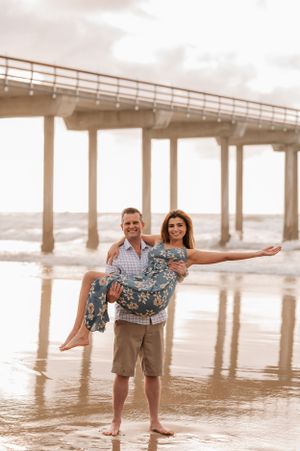Playful beach photo under pier with person carrying another in floral dress.