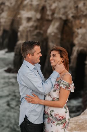 Romantic couple embracing on rocky coastal cliffs during sunset photoshoot in coordinated blue and floral outfits.