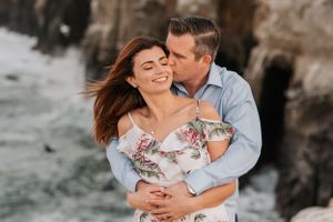 Couple sharing romantic moment on rocky beach cliffs during golden hour photography session.