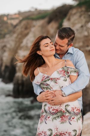 Loving couple in coordinated outfits dancing and embracing by ocean cliffs during sunset photoshoot.