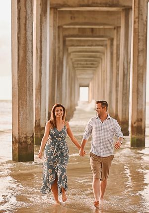 A couple splashing and playing in the water under a pier at sunset.