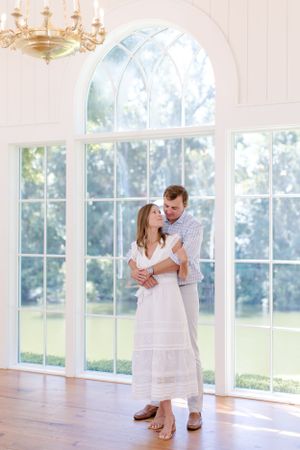 A romantic couple embraces in a sunlit room with large arched windows and hardwood floors.