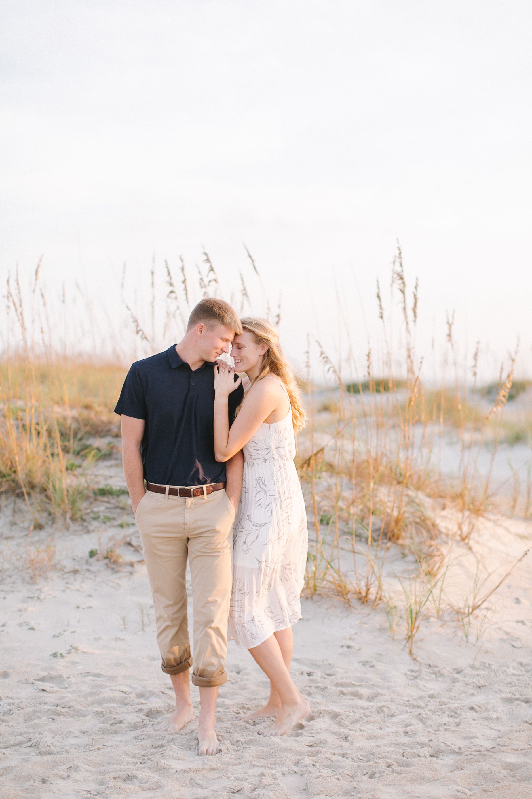 Romantic couple walking barefoot together on sandy beach dunes at sunset.