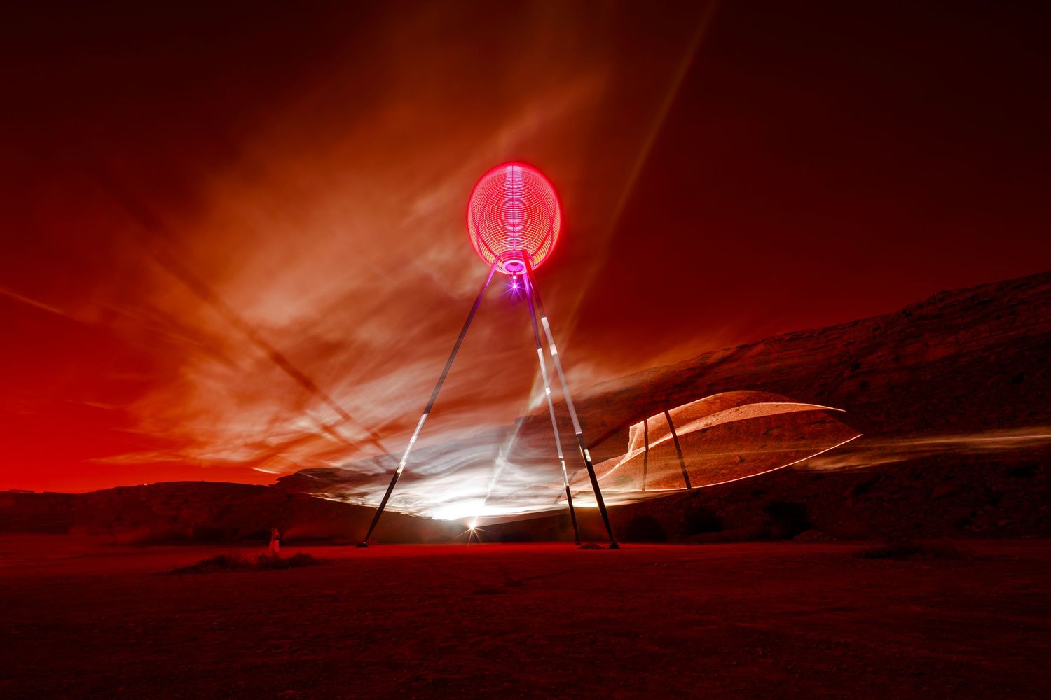 A giant red orb installation glows against a dramatic sunset sky in a desert landscape.