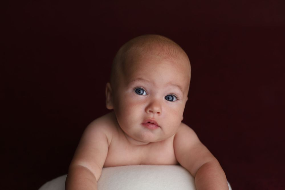 Alert baby with blue eyes poses on white surface against deep red background.
