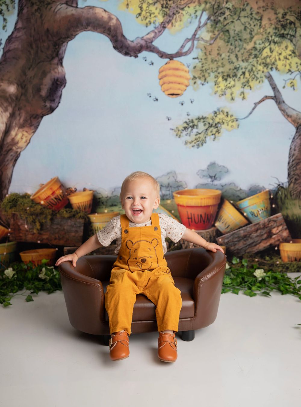 A laughing baby in Winnie the Pooh overalls poses in a leather chair with a Hundred Acre Wood backdrop.