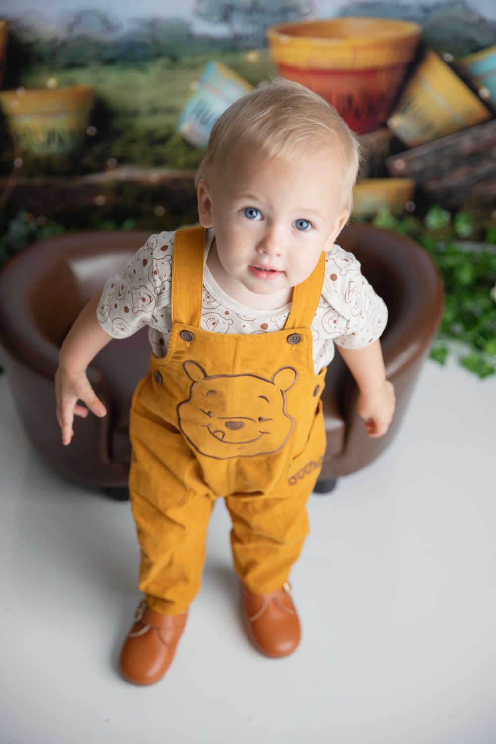 An adorable baby in Winnie the Pooh overalls stands in a Hundred Acre Wood themed photo studio.