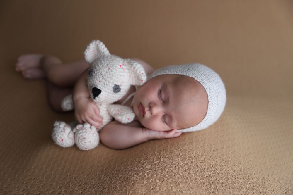 Sleeping newborn in white knit bonnet cuddles crocheted teddy bear on beige backdrop.