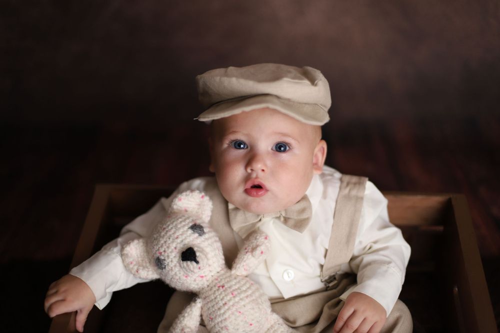 Blue-eyed baby in vintage newsboy cap and suspenders holds knit teddy bear in wooden crate.