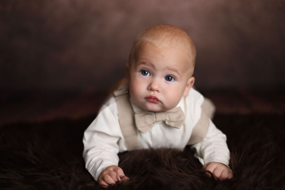 Blue-eyed baby in white outfit with bow tie poses on fluffy brown fur backdrop.