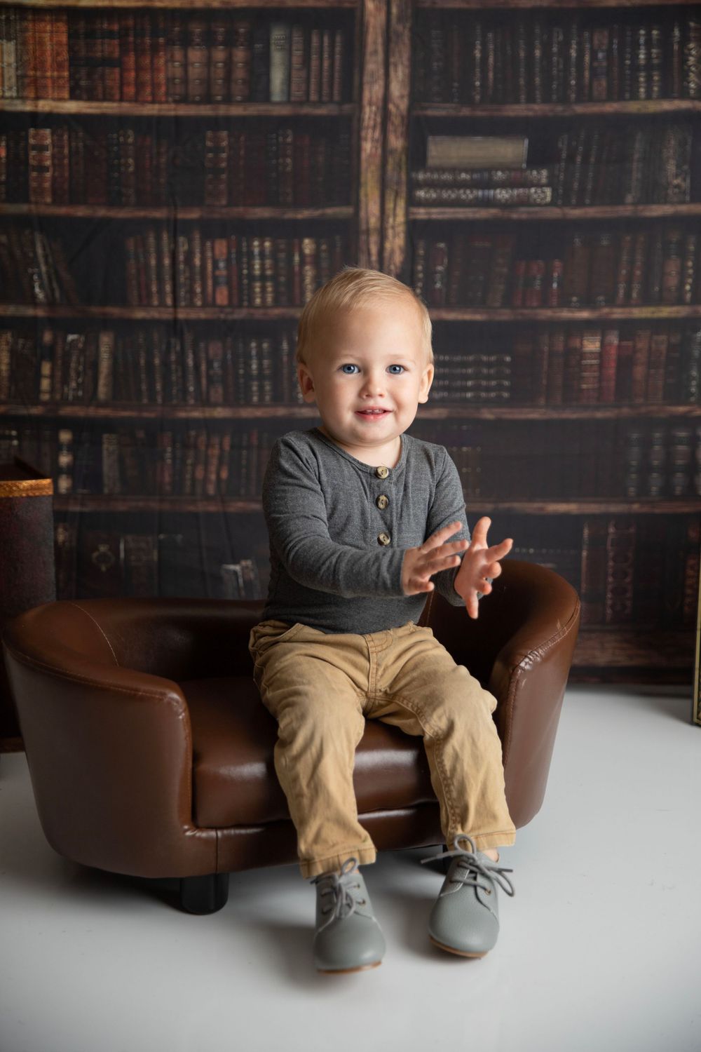 A happy toddler boy in khaki pants and gray shirt sits in a leather chair before a bookshelf backdrop.