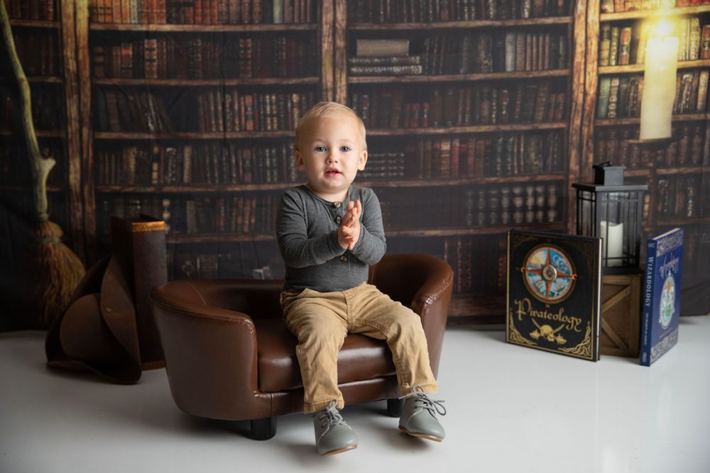 A cute toddler boy claps his hands while seated in a small leather chair in a library-themed studio.