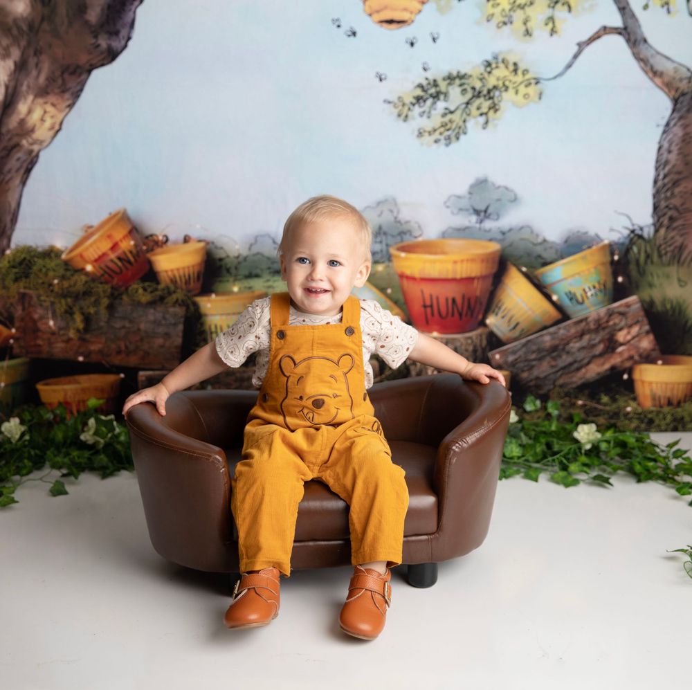 A joyful baby in yellow Winnie the Pooh overalls sits in a leather chair in a storybook themed studio.