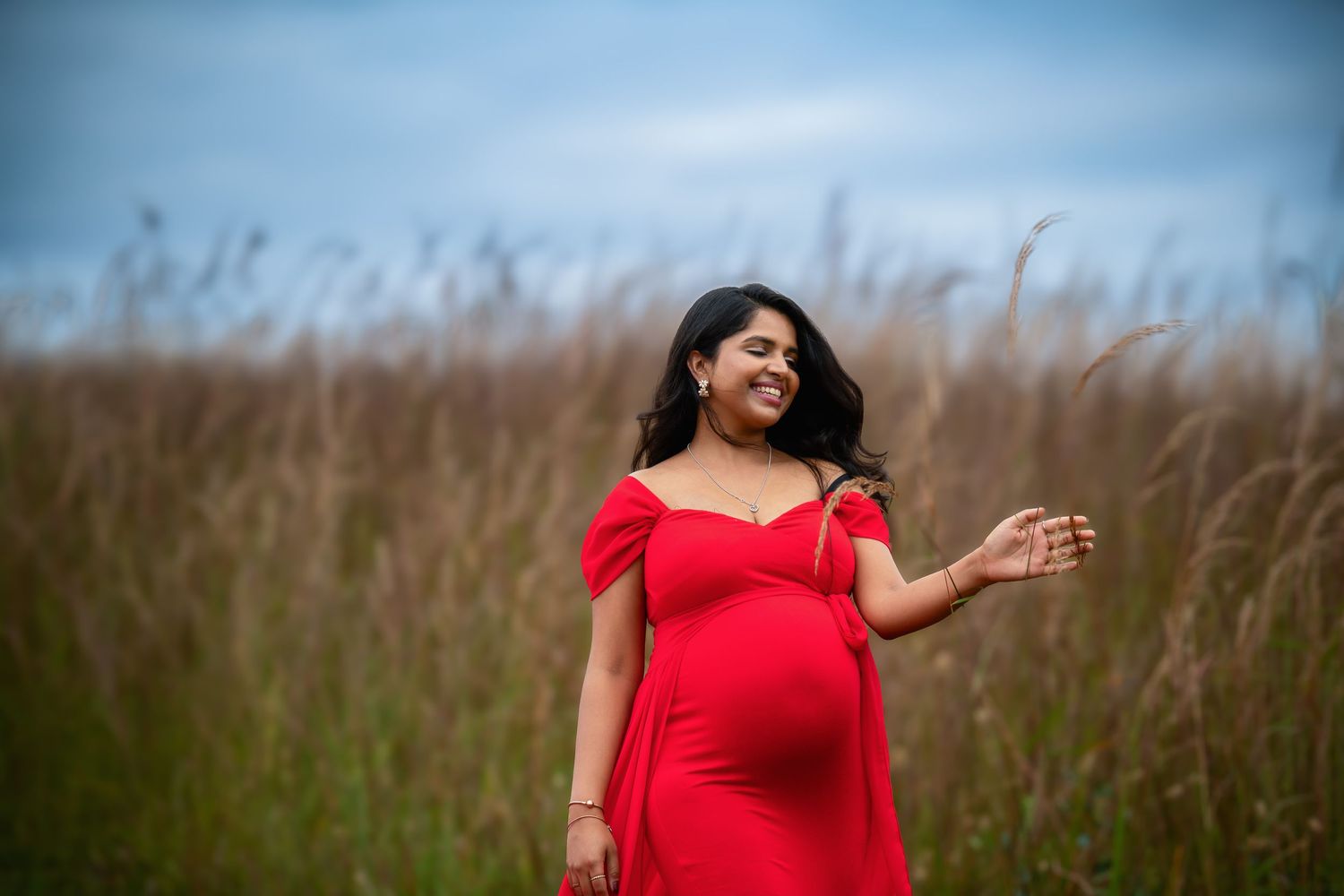 Person in red dress poses joyfully in field of tall grasses during maternity session.