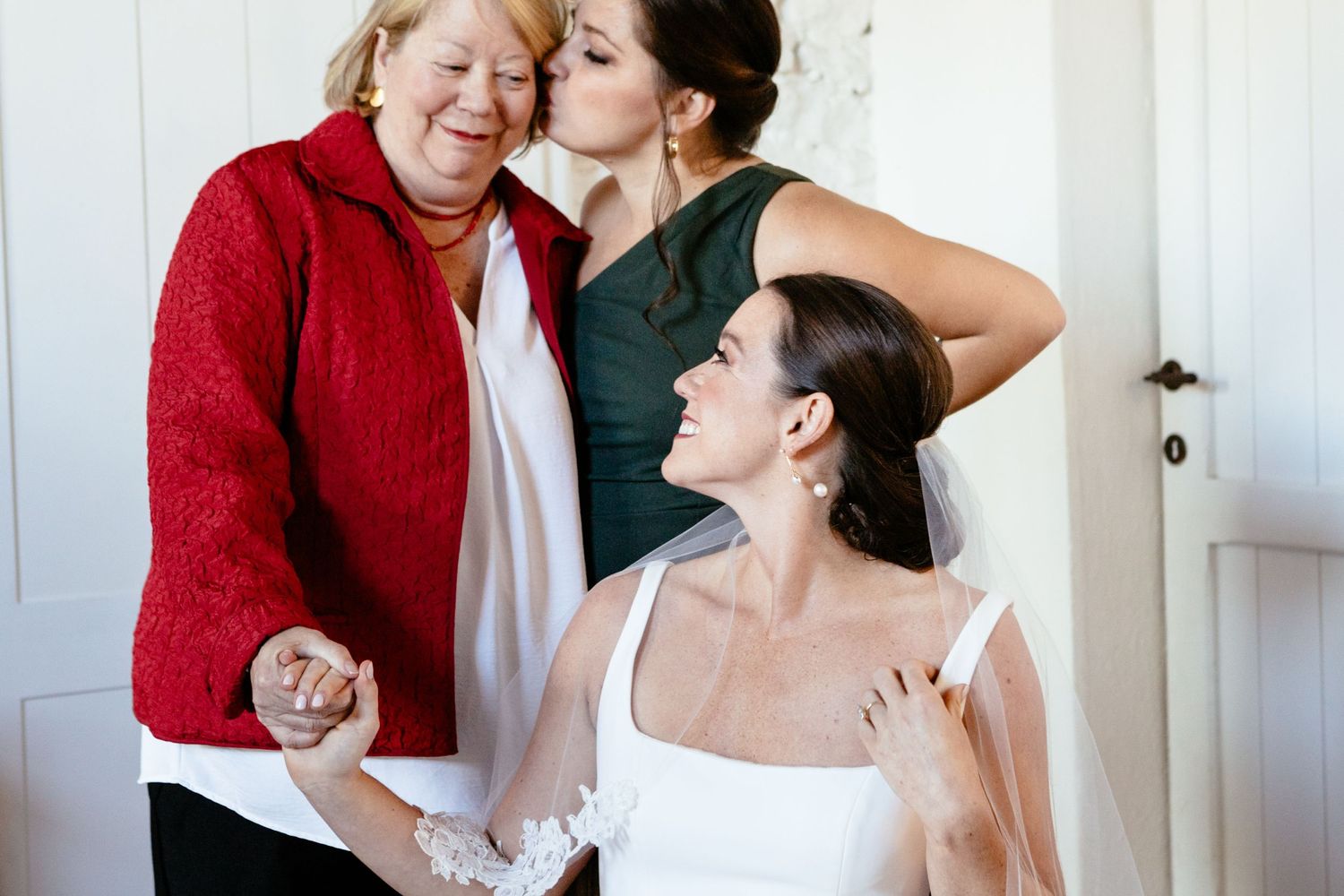The bride holds hands and looks at her mother while a bridesmaid gives her a kiss on the check.